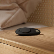 A We-Vibe Moxie + Black Hands-Free Remote or App Controlled Panty Vibrator sits on a wooden bedside table beside a bed with beige bedding and a wicker headboard.
