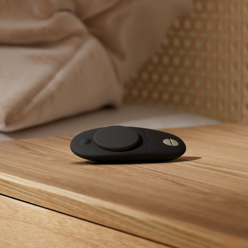 A We-Vibe Moxie + Black Hands-Free Remote or App Controlled Panty Vibrator sits on a wooden bedside table beside a bed with beige bedding and a wicker headboard.