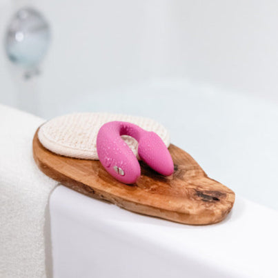 A We-Vibe Sync 2 Wearable Couples Vibrator in Dusty Pink and a textured round cloth rest on a wooden tray by the edge of a white bathtub.