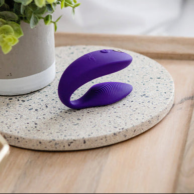 A purple We-Vibe Sync 2 Wearable Couples Vibrator, featuring a curved silicone design with ridges, rests on a round speckled stone tray beside a potted plant on a wooden surface.