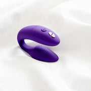 The We-Vibe Sync 2 Wearable Couples Vibrator, a purple remote and app-controlled silicone vibrator, features a ribbed texture and control buttons, resting on a white fabric surface.