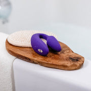 A We-Vibe Sync 2 Wearable Couples Vibrator (Remote and App Controlled, Purple) rests on a wooden tray beside a textured pad at the edge of a bathtub.
