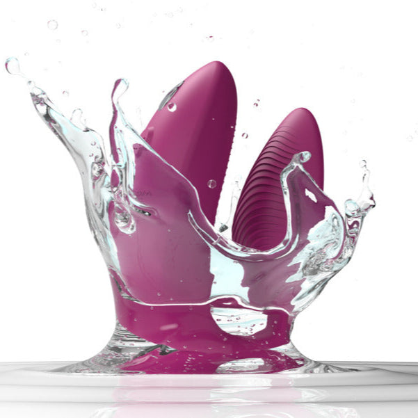 A dusty pink We-Vibe Sync 2 wearable couples vibrator, remote and app controlled, splashes into clear water, sending dynamic droplets and ripples across a white background.
