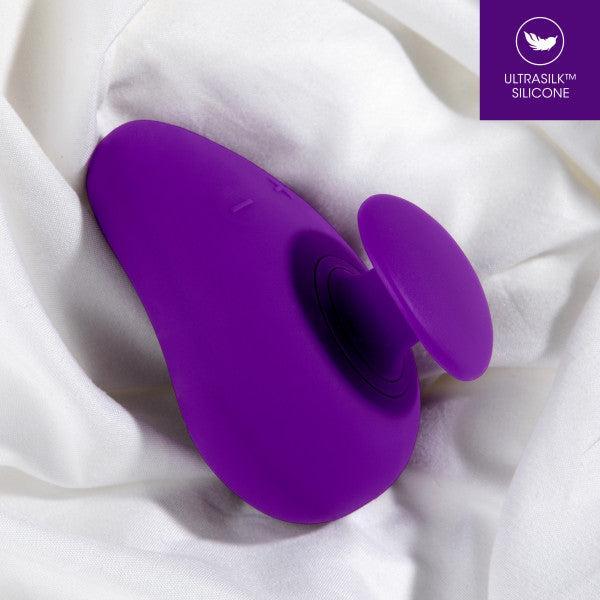 A purple Blush Wellness Palm Sense Rumbly External Vibrator with Finger Hold lies on a bed of white fabric. The ergonomic massager has a curved body and a round suction head. The top right corner features a label stating "ULTRASILK™ SILICONE" with a stylized bird logo.