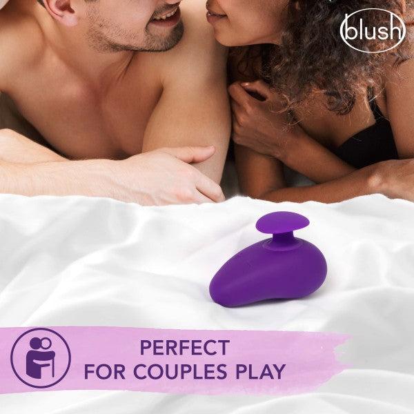 A couple lies in bed with a pink Wellness Palm Sense Rumbly External Vibrator with Finger Hold by Blush nearby. The image promotes intimacy with the text "blush" and "Perfect for Couples Play." Relaxed and smiling, the couple's focus is on each other while the toy is centrally placed on the white bedspread.