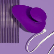 A purple, oval-shaped Wellness Palm Sense Rumbly External Vibrator with Finger Hold by Blush, featuring a circular attachment and a white cord, is placed against a matching purple background next to a ribbed, translucent silicone sheet. The overall scene is minimalistic and color-coordinated.