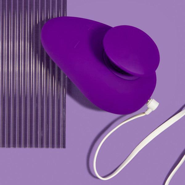 A purple, oval-shaped Wellness Palm Sense Rumbly External Vibrator with Finger Hold by Blush, featuring a circular attachment and a white cord, is placed against a matching purple background next to a ribbed, translucent silicone sheet. The overall scene is minimalistic and color-coordinated.