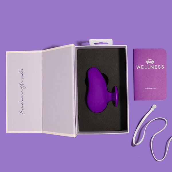 A purple, teardrop-shaped vibrator, identified as the Wellness Palm Sense Rumbly External Vibrator with Finger Hold by Blush, rests in a black foam insert within an open white box against a purple background. The box lid reads "Experience the vibe." Nearby, a purple booklet titled "Wellness" and a white USB charging cable accompany the silicone device.