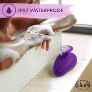 A close-up shot captures the Wellness Palm Sense Rumbly External Vibrator with Finger Hold, a purple waterproof rechargeable device from Blush, adorned with soapy bubbles and positioned on the edge of a bathtub. In the background, a person relaxes in a bubble bath. The text "IPX7 Waterproof" appears at the top alongside a water droplet icon, and the "Blush" logo is featured in the bottom right corner.