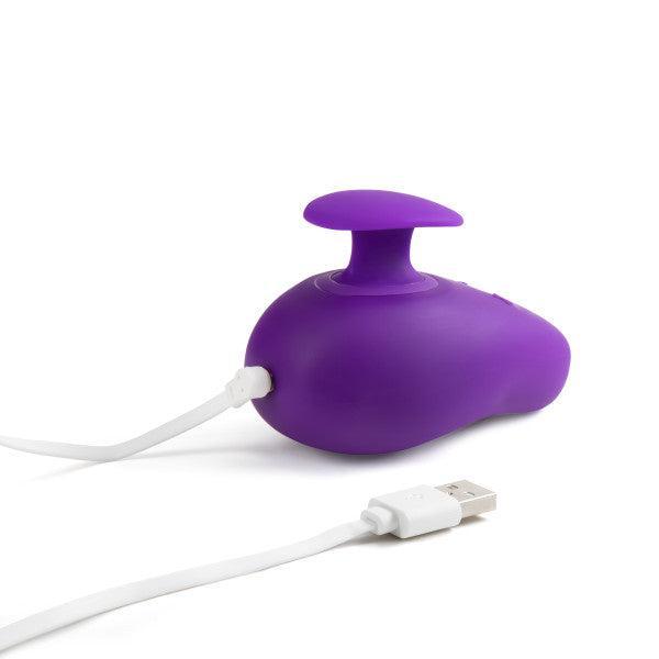The Wellness Palm Sense Rumbly External Vibrator with Finger Hold by Blush is a small purple silicone device with a rounded shape and a protruding button on top, connected to a white USB charging cable. The USB connector is lying next to the ergonomic massager.