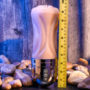 The Tight Lipped Vibrating Vanilla Stroker with Suction + Free Movie Download, featuring a flesh-toned design and control buttons on a metallic base, stands upright near rocks and a measuring tape, set against a wooden backdrop.