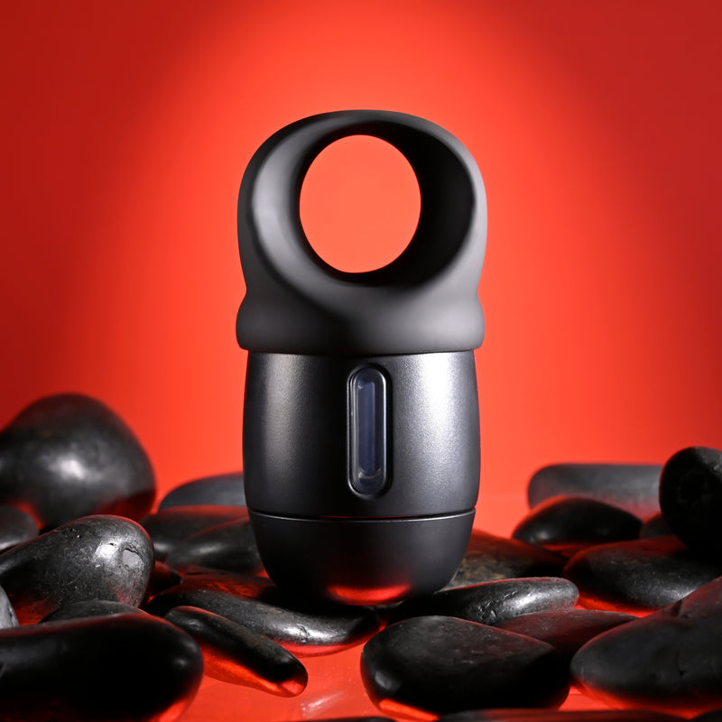 The Wet It Be Hand Held Vibrating Masturbator with Lube Dispenser, black and cylindrical with a large circular handle, sits among smooth black stones against a vibrant red background.