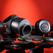 The Wet It Be Hand Held Vibrating Masturbator with Lube Dispenser, in matte black with "PUKP" branding on both bottle and cap, lies open on smooth black stones against a vibrant red backdrop, showcasing its sleek modern design.