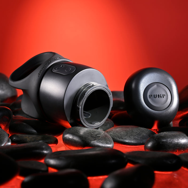 The Wet It Be Hand Held Vibrating Masturbator with Lube Dispenser, in matte black with "PUKP" branding on both bottle and cap, lies open on smooth black stones against a vibrant red backdrop, showcasing its sleek modern design.