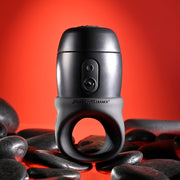 The Wet It Be Hand Held Vibrating Masturbator with Lube Dispenser, in black and gray with buttons, is shown upright among smooth black stones on a red background.