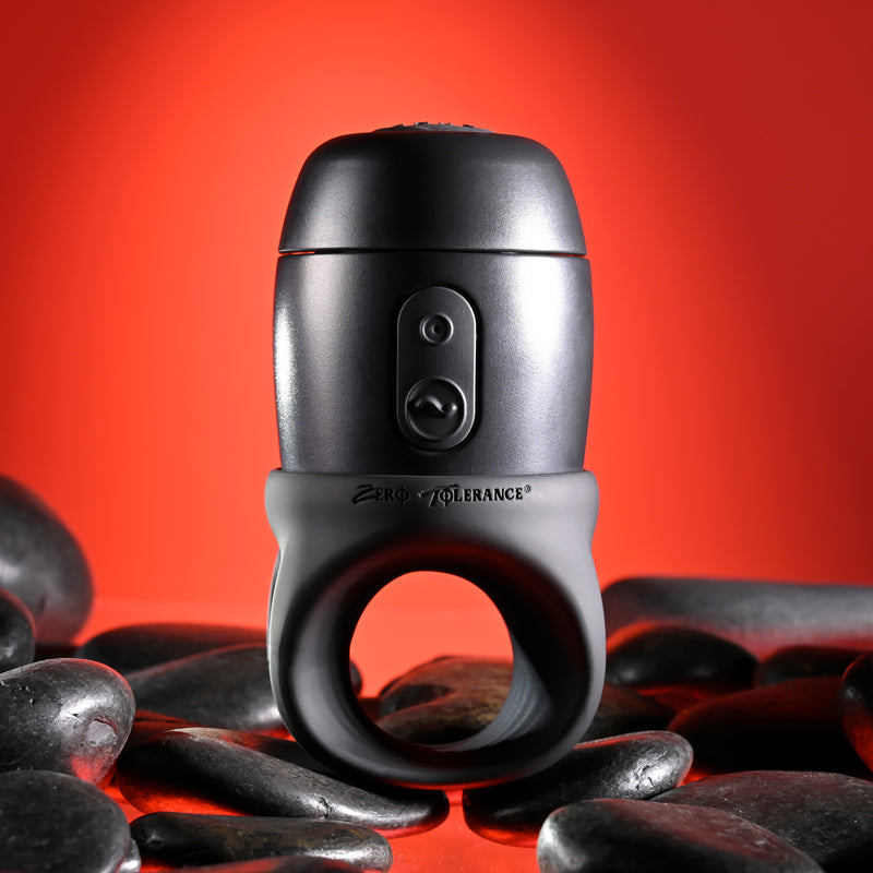 The Wet It Be Hand Held Vibrating Masturbator with Lube Dispenser, in black and gray with buttons, is shown upright among smooth black stones on a red background.
