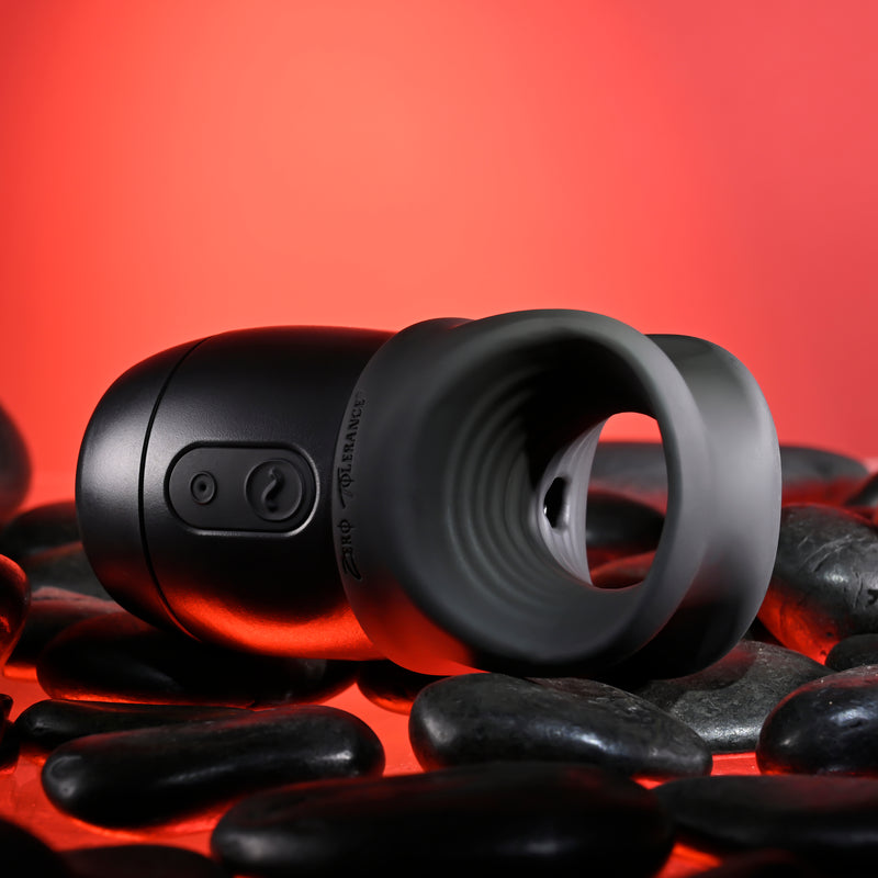The Wet It Be Hand Held Vibrating Masturbator with Lube Dispenser, in black and gray with a rounded, open-ended design, rests on smooth black stones against a red background.