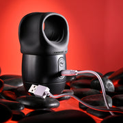 The Wet It Be Hand Held Vibrating Masturbator with Lube Dispenser, featuring a black design and circular opening, is shown charging via USB cable on smooth black stones with a red background.