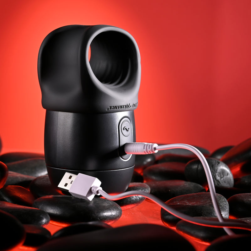 The Wet It Be Hand Held Vibrating Masturbator with Lube Dispenser, featuring a black design and circular opening, is shown charging via USB cable on smooth black stones with a red background.
