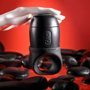 A white mannequin hand holds the Wet It Be Hand Held Vibrating Masturbator with Lube Dispenser, featuring buttons and a ring base, displayed on smooth black stones with a red background.