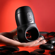 A hand holds the Wet It Be Hand Held Vibrating Masturbator with Lube Dispenser, featuring front buttons, surrounded by dark smooth stones against a red background.