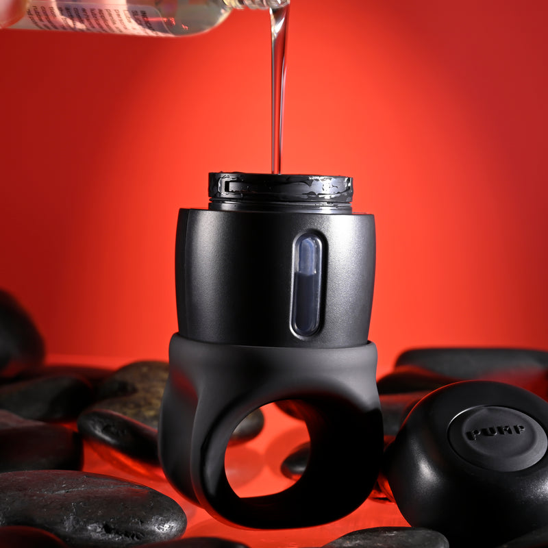 A close-up of the Wet It Be Hand Held Vibrating Masturbator with Lube Dispenser, shown among smooth black stones. Liquid is being poured into its top opening against a red background, with a matching black cap placed beside it.