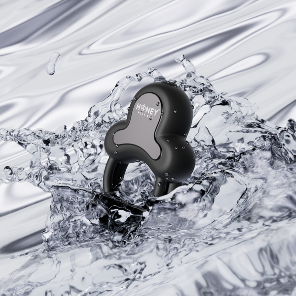 The Anello App-Controlled Vibrating Double Cock Ring with Clitoral Stimulator, in black and marked "HONEY PLAY BOX", features dual silicone rings splashing into clear water against a shiny, metallic background.