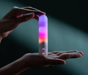 Hands holding a white, sleek bullet vibrator glowing with purple, pink, and orange LED lights.