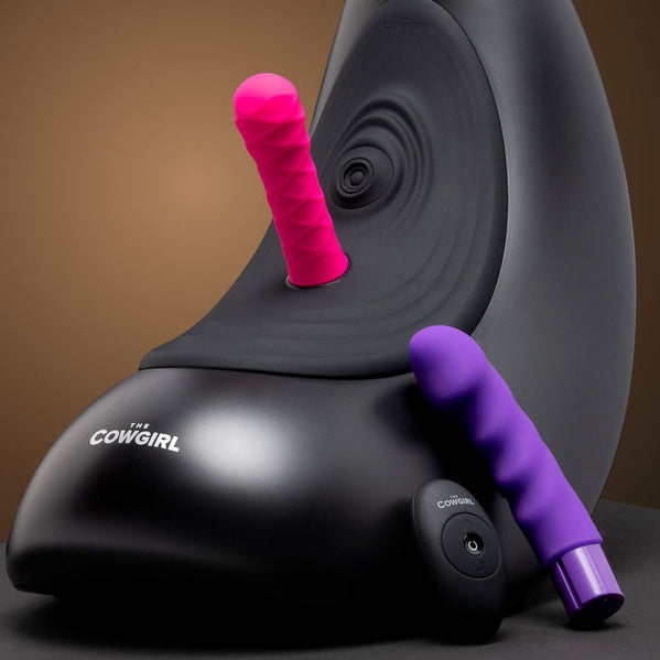 The Cowgirl Saddle Sex Machine with Vibration, Thrusting and Rocking is shown in black with a pink textured attachment on top and a purple one beside it, offering hands-free pleasure against a brown backdrop.