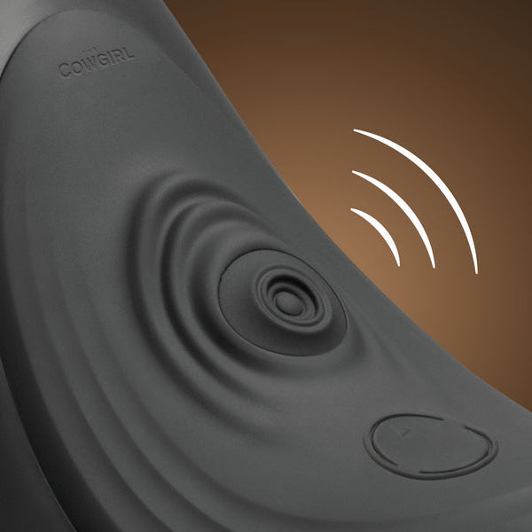 Close-up of The Cowgirl Saddle Sex Machine with Vibration, Thrusting and Rocking in black, featuring ripple patterns, a round button, and "COWGIRL" engraving; white lines evoke hands-free pleasure and vibration on a brown background.
