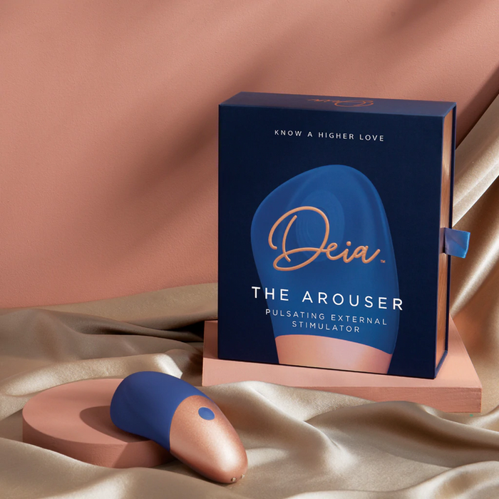 A blue and rose gold The Arouser Pulsating External Stimulator, made from body-safe silicone, is displayed on satin fabric beside its matching box labeled "Deia The Arouser Pulsating External Stimulator" in gold and white letters.