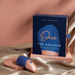 A blue and rose gold The Arouser Pulsating External Stimulator, made from body-safe silicone, is displayed on satin fabric beside its matching box labeled "Deia The Arouser Pulsating External Stimulator" in gold and white letters.