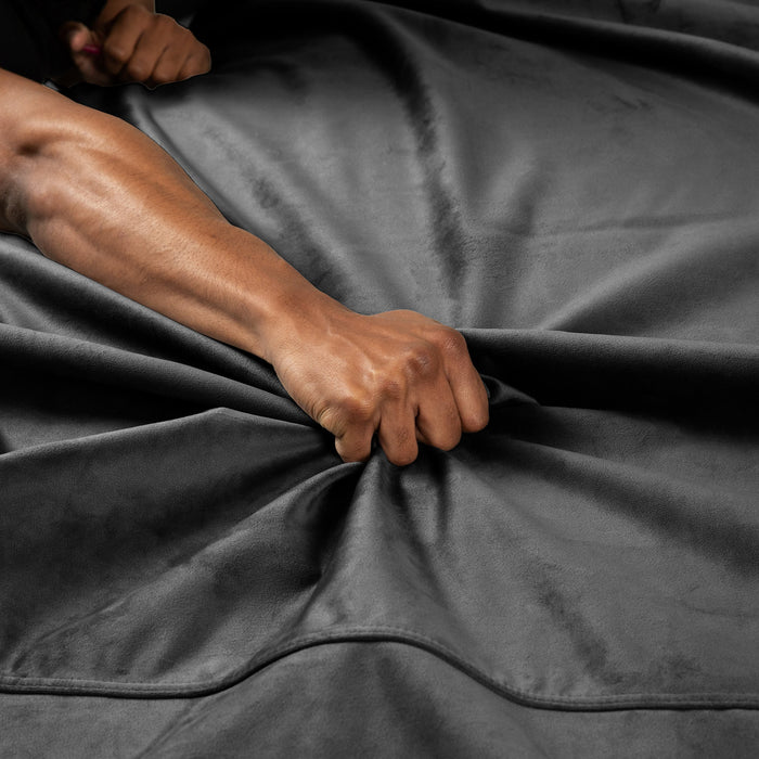 A close-up shows a hand gripping and pulling the dark, velvety Liberator Fascinator Throw Waterproof Sex Blanket, creating visible folds and creases. Another hand appears partially in the background.