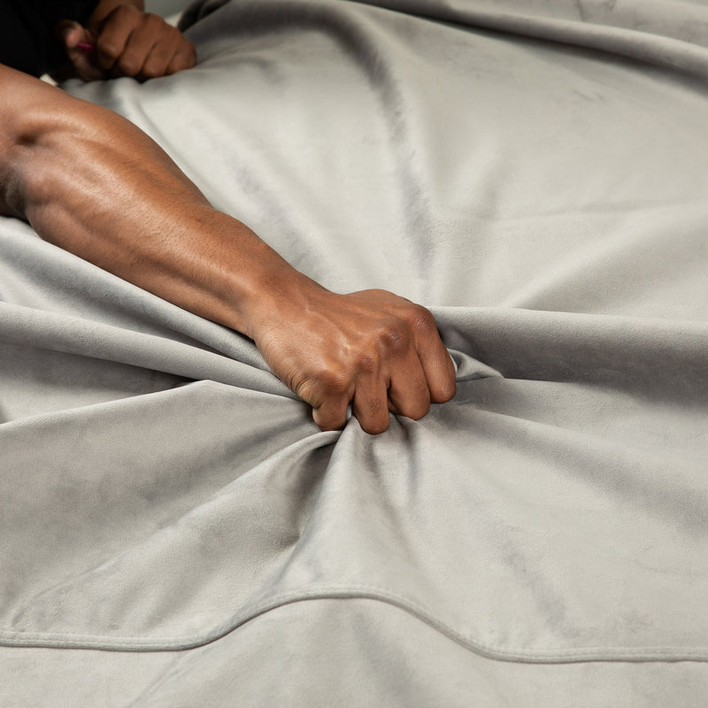 A hand grips and gathers the soft, light gray Liberator Fascinator Throw Velvety Waterproof Sex Blanket, forming visible folds and tension in the fabric.