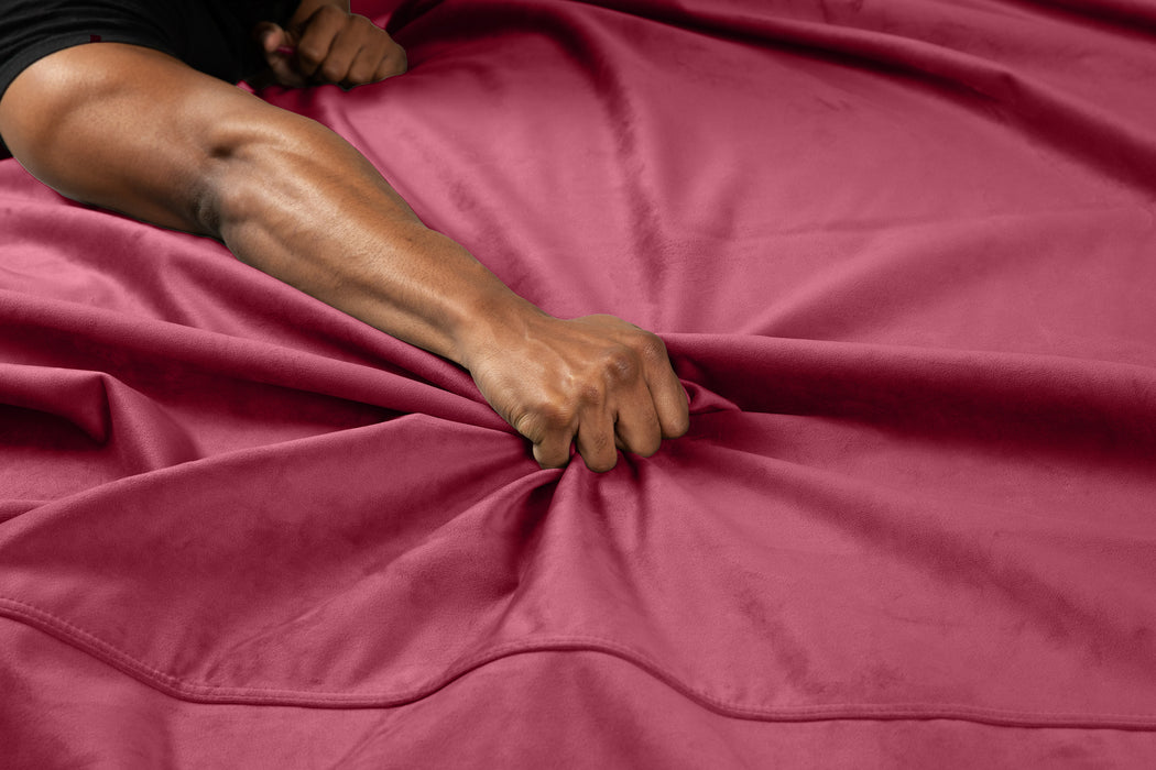 A close-up of a hand gripping and bunching up the red Liberator Fascinator Throw Velvety Waterproof Sex Blanket on a bed, with an extended arm and visible muscles.