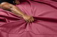 A close-up of a hand gripping and bunching up the red Liberator Fascinator Throw Velvety Waterproof Sex Blanket on a bed, with an extended arm and visible muscles.