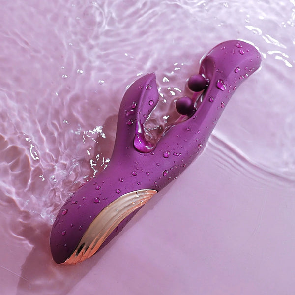 The Gem Pressure Sensing App-Controlled Twin Balls Rabbit (Damaged Box), a purple waterproof personal massager with gold accents, rests on a wet light pink surface surrounded by water droplets and gentle ripples.