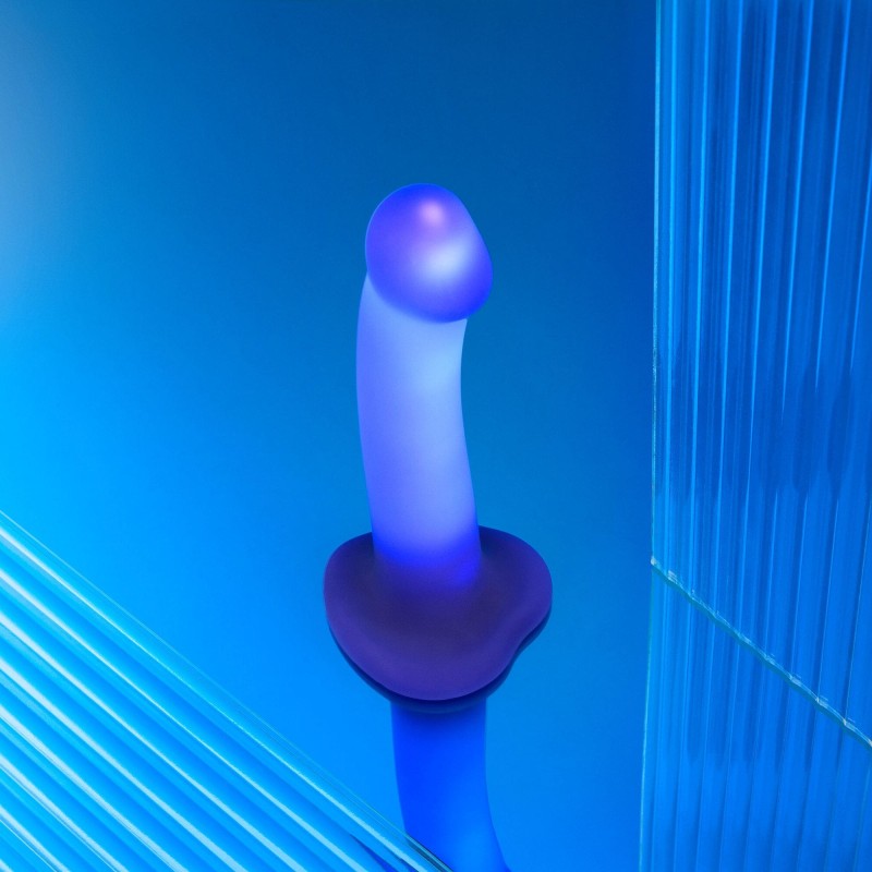 The Glow Blue LED 7 Inch Light Up Silicone Dildo, featuring a flared base and strap-on compatibility, stands upright on a reflective blue surface amid vertical and diagonal ridged acrylic panels.