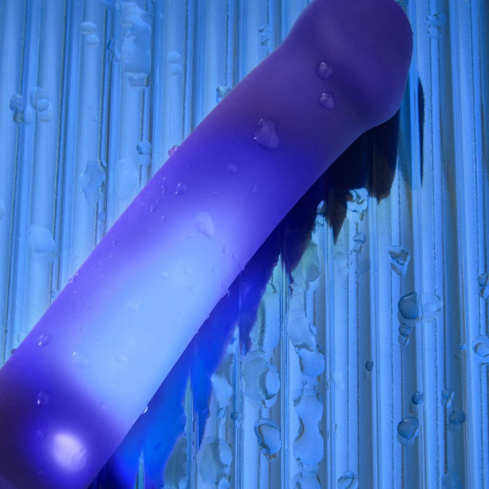 The Glow Blue LED 7 Inch Light Up Silicone Dildo, accented with water droplets, rests on a blue ribbed reflective surface—its glowing blue LED feature enhancing the vibrant and glossy appearance.