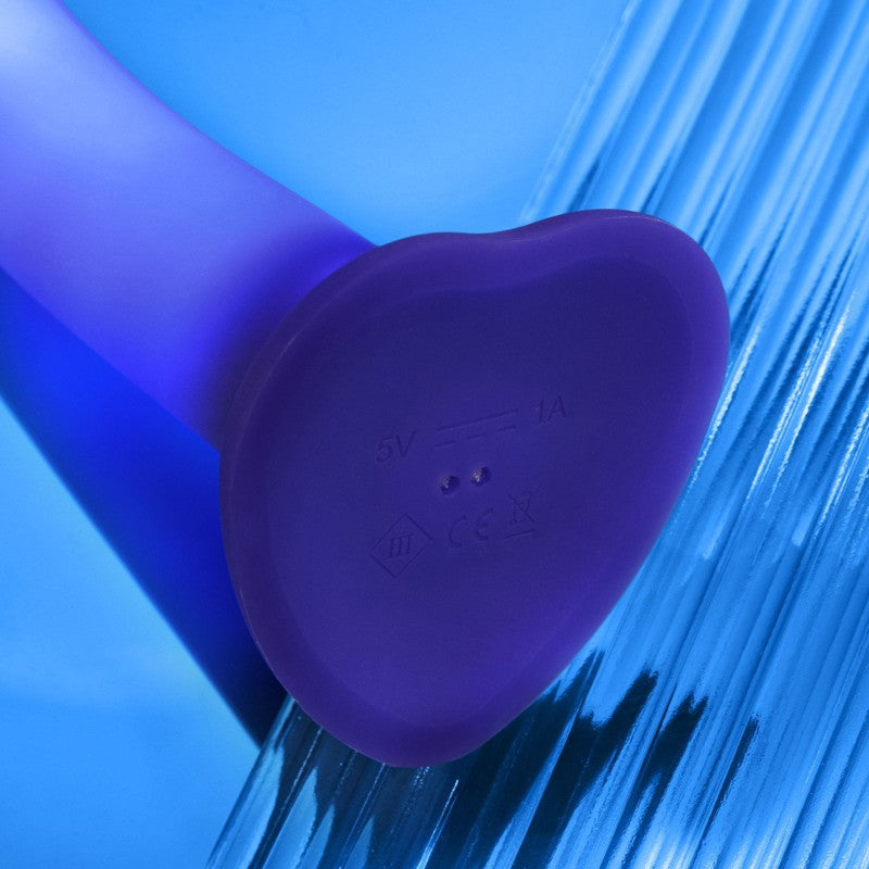 Close-up of the Glow Blue LED 7 Inch Light Up Silicone Dildo’s heart-shaped base with charging icons and regulatory markings, set on a blue textured background. This purple silicone toy is harness compatible for versatile strap-on use.