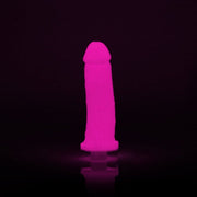 The Clone A Willy Vibrating Silicone Penis Casting Kit in Glow In the Dark Pink stands upright, casting a pink glow against a dark background.