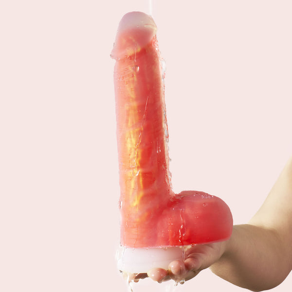 A hand holds the Noctura Thrusting Silicone Dildo with App Control, a large pink and white realistic liquid silicone dildo, against a pale pink background as water is poured over it.