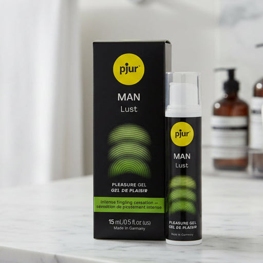 A bottle of pjur MAN Lust Penis Stimulating Gel stands next to its box on a bathroom counter. The black packaging displays green and white text and graphics, with bathroom items and a sink blurred in the background.