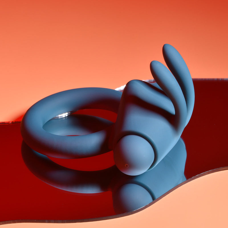 The Playboy Bunny Buzzer Vibrating Silicone Cock Ring, made from body-safe silicone with three curved extensions, is displayed on a reflective wavy red surface set against a gradient orange background.