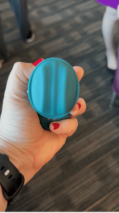 A hand with red-painted nails holds the Riana Circular Clitoral Rubbing Vibrator, made of body-safe teal silicone. The person wears a black watch, and the carpeted floor and parts of people's legs are visible in the background.