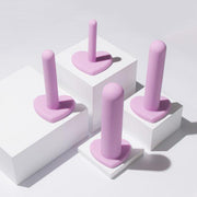 Four pastel pink silicone tools with heart-shaped bases are displayed on white platforms with shadows. The Wellness Vaginal Dilator Kit Graduated Set of 4 by Blush features cylindrical, upright tools crafted from body-safe silicone, arranged creatively under soft lighting.