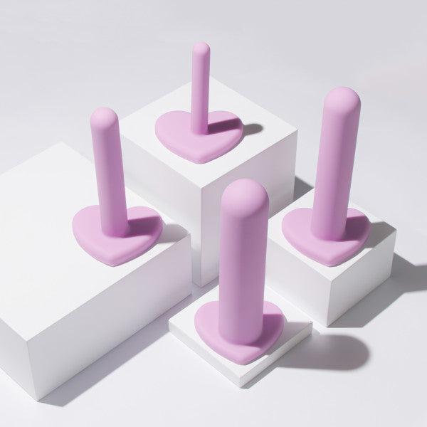 Four pastel pink silicone tools with heart-shaped bases are displayed on white platforms with shadows. The Wellness Vaginal Dilator Kit Graduated Set of 4 by Blush features cylindrical, upright tools crafted from body-safe silicone, arranged creatively under soft lighting.