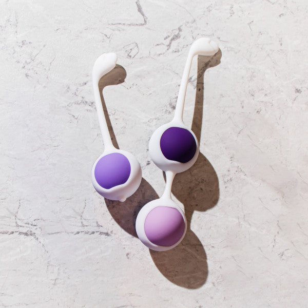 The Wellness Kegel Ball Training Kit for Pelvic Floor Health features two silicone Kegel balls with long handles in purple and pink, lying side by side on a light marble surface—ideal for effective pelvic floor strengthening.