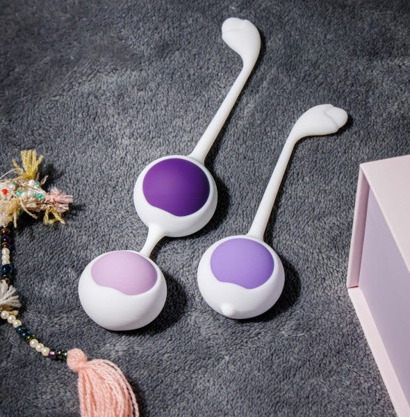 The Wellness Kegel Ball Training Kit for Pelvic Floor Health includes two sets of Kegel balls with white handles and purple accents, displayed on a soft gray surface beside a beaded tassel and a pink box—ideal for pelvic floor strengthening.